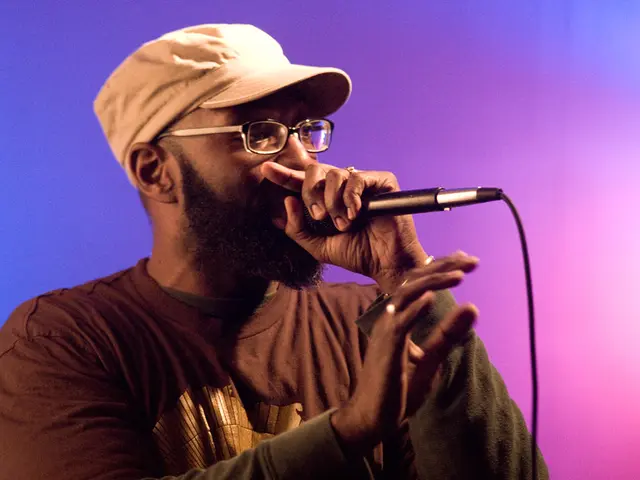 One man is standing and wearing a brown t-shirt and wearing glasses and a cap, he is talking in the...